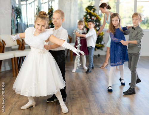 Couple of smiling tweens, boy and girl in festive clothing performing graceful waltz with group classmates under guidance of female teacher during Christmas event at school..