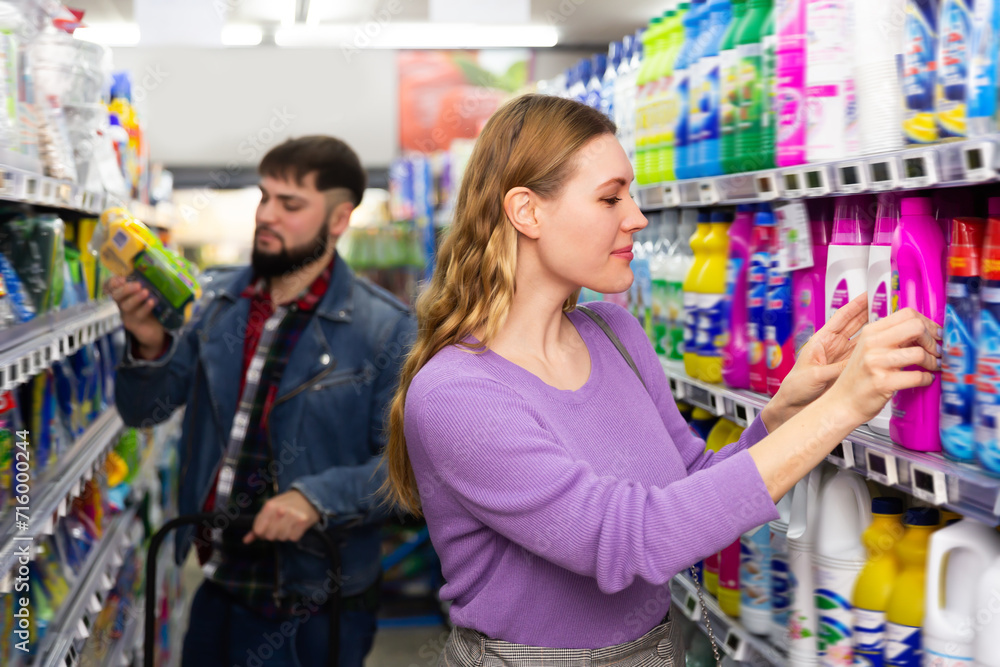 Young people making purchases in household chemistry store, woman choosing detergent, man on background