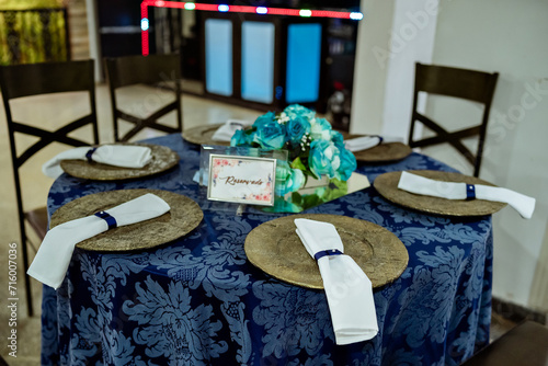 a round table decorated with a blue tablecloth, plate holder and a flower arrangement with a glass vase