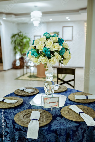 a round table decorated with a blue tablecloth, plate holder and a flower arrangement with a glass vase