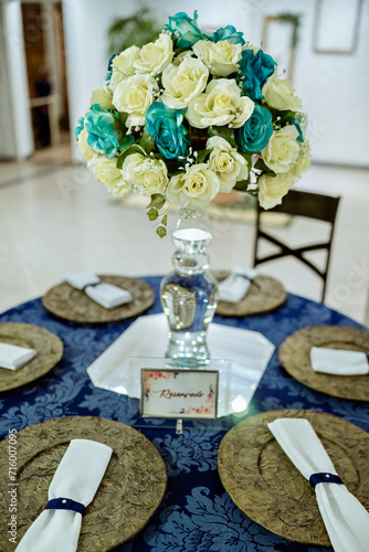 a round table decorated with a blue tablecloth, plate holder and a flower arrangement with a glass vase