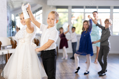 Enthusiastic preteen children, boys and girls in party attire performing elegant waltz in pairs in sunny hall of dance school with smiling female teacher in background..