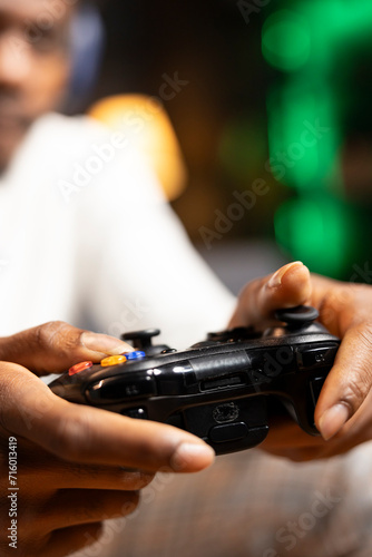 Focus shot on controller held by gamer in blurry background playing videogames. Close up shot of gamepad used by man enjoying gaming session in living room while sitting down in front of TV