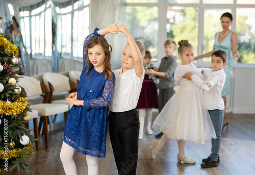 Enthusiastic preteen children, girls and boys in elegant outfits practicing partner dance moves in studio adorned with Christmas decorations, under guidance of female teacher