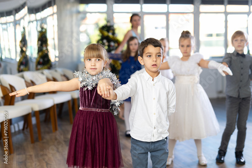 Enthusiastic preteen children, girls and boys in elegant outfits practicing partner dance moves in studio adorned with Christmas decorations, under guidance of female teacher