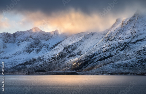 Snowy mountains in low clouds, sea bay, reflection in water at sunset in winter. Lofoten islands, Norway. Colorful landscape with rocks in snow, orange sky. Seashore. Nature background. Wintry scenery