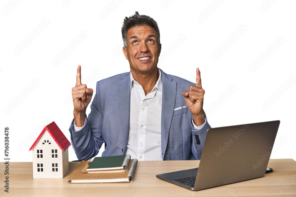 Middle-aged Hispanic businessman with real estate theme pointing upside with opened mouth. Middle-aged Hispanic businessman with real estate theme pointing upside with opened mouth.