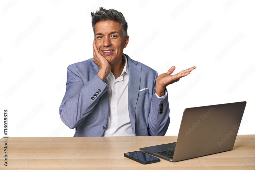 Elegant businessman at desk with laptop holds copy space on a palm, keep hand over cheek. Amazed and delighted. Elegant businessman at desk with laptop holds copy space on a palm, keep hand over cheek. Amazed and delighted.