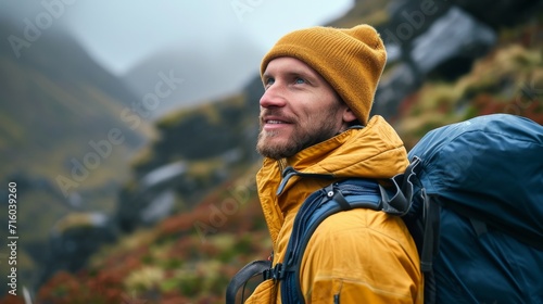 A rugged man with a sunny disposition stands confidently against a breathtaking mountain backdrop, donning a yellow hat and backpack while dressed for outdoor adventure in a warm coat and jacket