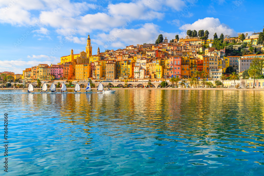 A small group of sailboats pass in front the old town and Les Sablettes Beach and promenade along the Cote d'Azur French Riviera at Menton, France.