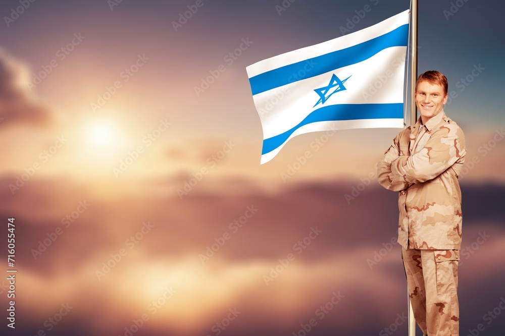 Soldier in uniform with Israeli flag against a sky.