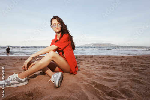 Cheerful woman sitting on a sandy beach, enjoying a sunny vacation day by the ocean - A carefree portrait of a trendy young girl with a fashionable outfit and a beautiful tan, radiating happiness and