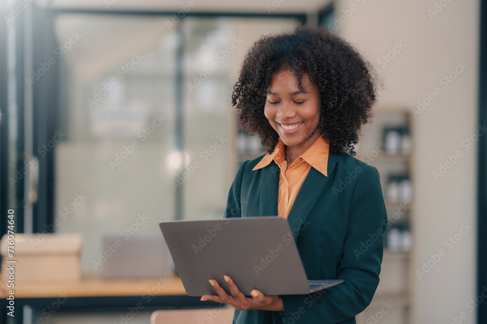 Smiling woman with curly hair holds a laptop, wearing a green blazer and orange blouse, standing in a modern office. Smiling woman with curly hair holds a laptop, wearing a green blazer and orange blouse, standing in a modern office.