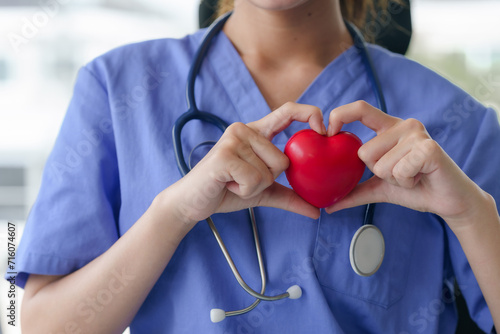 A nurse in blue scrubs holds a red heart in her hands, forming a heart shape around it, symbolizing care. photo