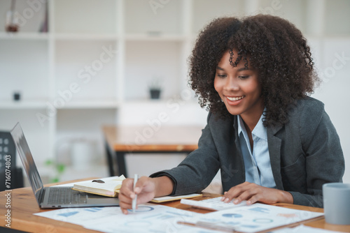 Smiling young professional woman reviewing financial documents at her workplace with laptop and coffee cup. photo