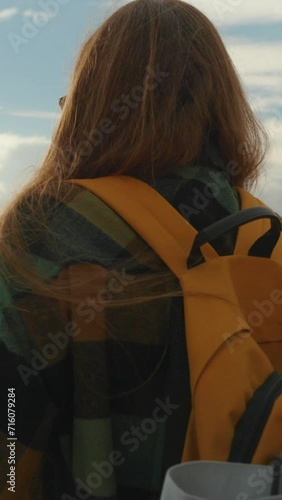 Vertical video. At the mountain summit, a woman watches the sunset, a backpack behind her, and her hair billowing in the wind. In the distance, the sea and mountains with dense forests. She enjoys tra photo