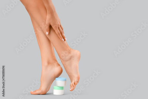 Beautiful young woman with jars of cream making foot massage on grey background