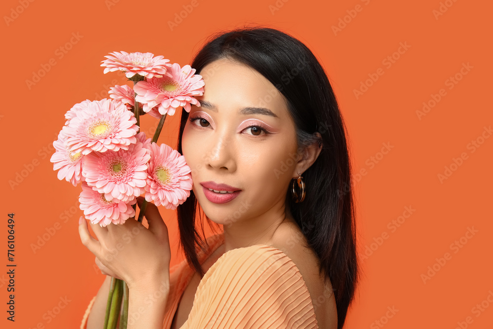 Fototapeta premium Beautiful young Asian woman with bouquet of pink gerbera flowers on orange background