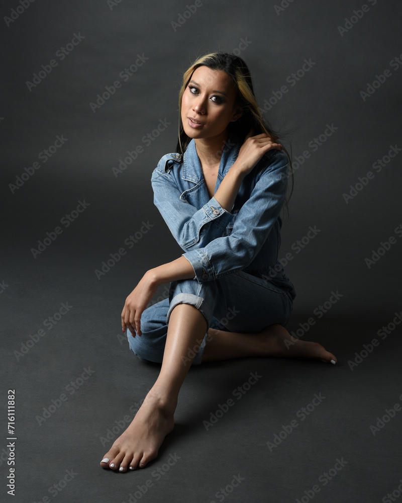Full length portrait of brunette female asian model wearing casual clothes, double denim jean pants. Isolated on dark studio background with shadows