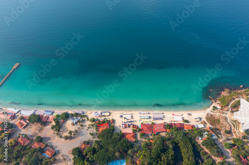 Aerial top view of colorful umbrellas on sandy beach, swim in blue sea summer sunny day. Tropical island sandy azure bay pier village buildings settlement surrounded exotic green forests trees hills.