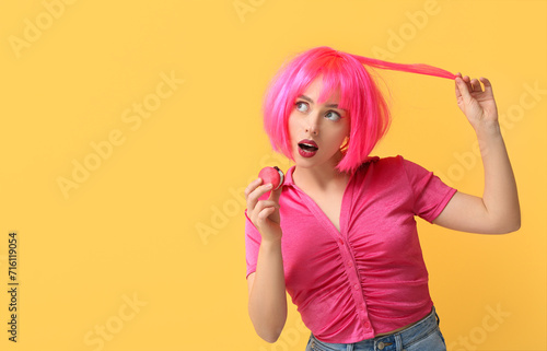 Beautiful young woman in pink wig eating sweet macaroon on yellow background