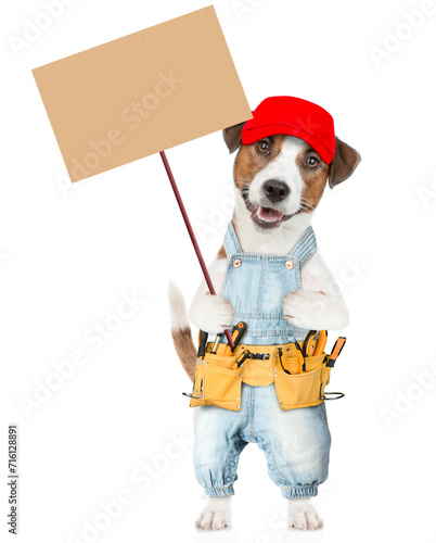Funny Jack russell terrier puppy wearing red cap and denim overalls with tool belt holds adjustable wrench and shows empty placard. isolated on white background