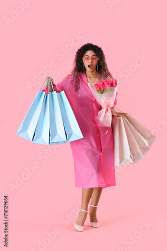 Fashionable African-American woman with bouquet of flowers and shopping bags on pink background. International Women's Day celebration