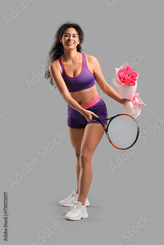 Young African-American woman in sportswear with bouquet of flowers and tennis racket on grey background. International Women's Day celebration © Pixel-Shot