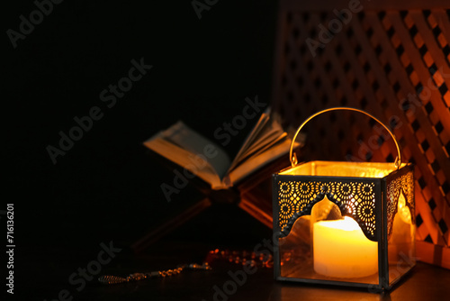 Muslim lantern with burning candle and Koran for Ramadan on table against dark background