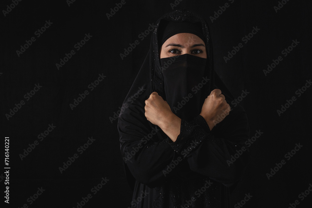 Portrait of Muslim female in black Hijab or Nigab showing stop gesture with crossed hands, over black background
