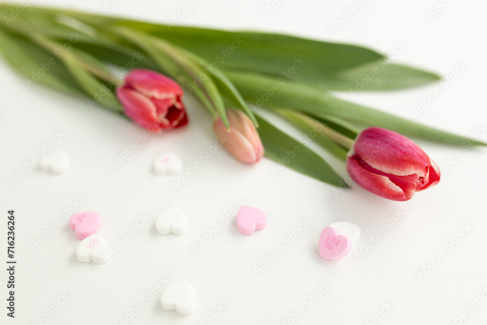 Fototapeta premium Background for Valentine's Day. Assorted pink, purple, white heart shaped candies with printed messages of love and fresh pink tulips on white background. Gift for Women's Day, March 8