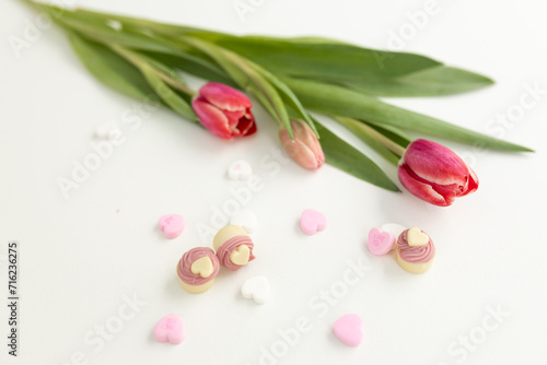 Fototapeta Naklejka Na Ścianę i Meble -  Background for Valentine's Day. Assorted pink, purple, white heart shaped candies with printed messages of love and fresh pink tulips on white background. Gift for Women's Day, March 8