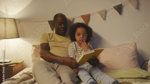 Medium shot of African American father reading fairy tale to little bored and uninterested boy in pajamas before sleep in modern cozy bedroom photo
