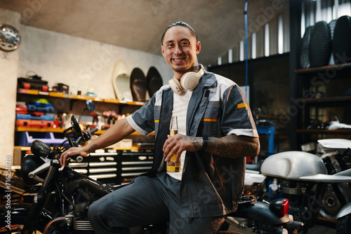 Portrait of happy mechanic with bottle of beer sitting on motorcycle he fixed for client photo