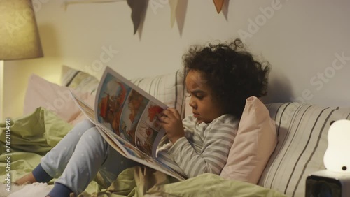 Side medium shot of little African American 5 year old boy in pajamas paging though comic strip about dinosaurs looking at cartoons lying in bed in evening photo
