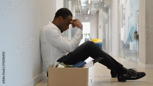 Fired black guy sitting frustrated and upset in the hallway near office with dismissal box photo