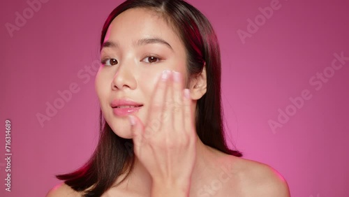 Young Asian girl applying cosmetic cream on face against pink background