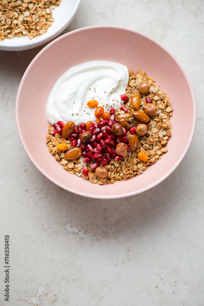 Roseate bowl with granola, greek yogurt, pomegranate and nuts, vertical shot on a light-beige stone background, elevated view Roseate bowl with granola, greek yogurt, pomegranate and nuts, vertical shot on a light-beige stone background, elevated view