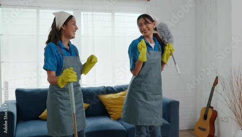Occupied with housework two women in uniforms stand in a dirty room using professional cleaning equipment. With smiles they work as a team cleaning furniture and ensuring living room purity. photo