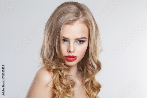 Charming young woman with clean fresh skin and long curly hair looking at camera on white studio wall background, portrait