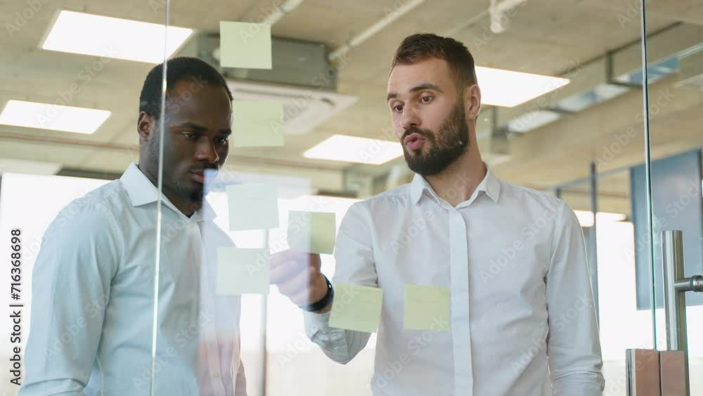 Two businessmen in meeting brainstorming and discussing post it notes stuck on glass wall at office Two businessmen in meeting brainstorming and discussing post it notes stuck on glass wall at office