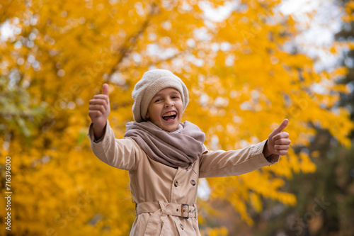 Happy caucasian girl in a beige coat and beret walks in the park in autumn. Schoolgirl showing thumbs up. 