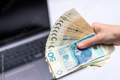 Plik polskich banknotów gotówki pln trzymany w dłoni, w tle komputer 