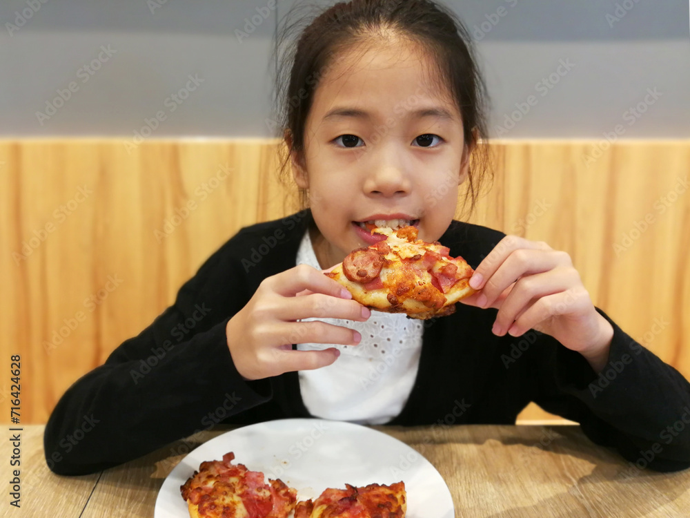 Happy Asian child enjoy eating pizza on table. Portrait of a young girl with an appetizing piece of pizza. Thai girl eating pizza at cafe, concept for unhealthy junk food. Focus on pizza. Happy Asian child enjoy eating pizza on table. Portrait of a young girl with an appetizing piece of pizza. Thai girl eating pizza at cafe, concept for unhealthy junk food. Focus on pizza.