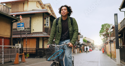 Bicycle, cycling and man by buildings for travel, exercise or transportation to college academy. Fitness, backpack and young male student driving a bike to university for carbon footprint by houses. photo