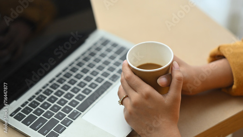 Cropped shot young woman holding coffee cup and using laptop on wooden desk
