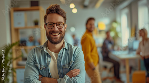 Confident Male Entrepreneur at Startup Office. A confident young male entrepreneur with glasses stands proudly in a bustling startup office environment.