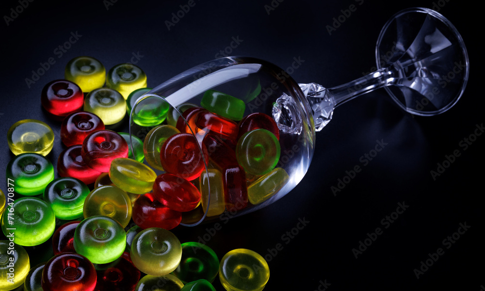 Fototapeta premium Many lollipops spill out from an overturned glass goblet. Multi-colored candies in a glass on a black background.