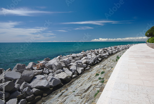 View of the blue sky with white clouds far in the background, the turquoise sea and the contrasting white, wide empty promenade in the tourist town of Grado, Italy.
