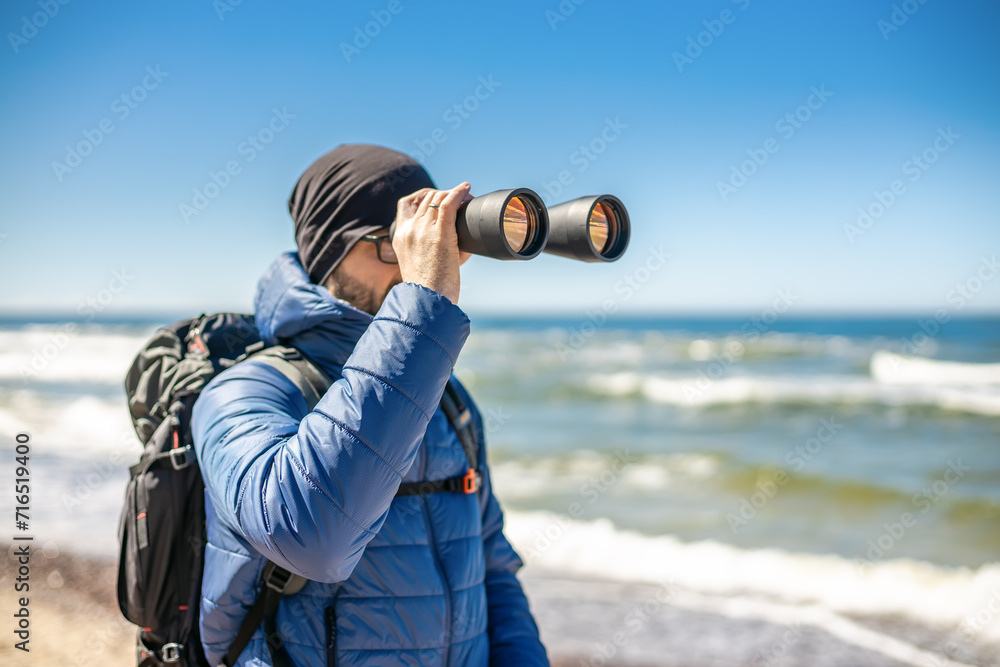 Obraz premium A tourist man standing by the seashore, looking through binoculars